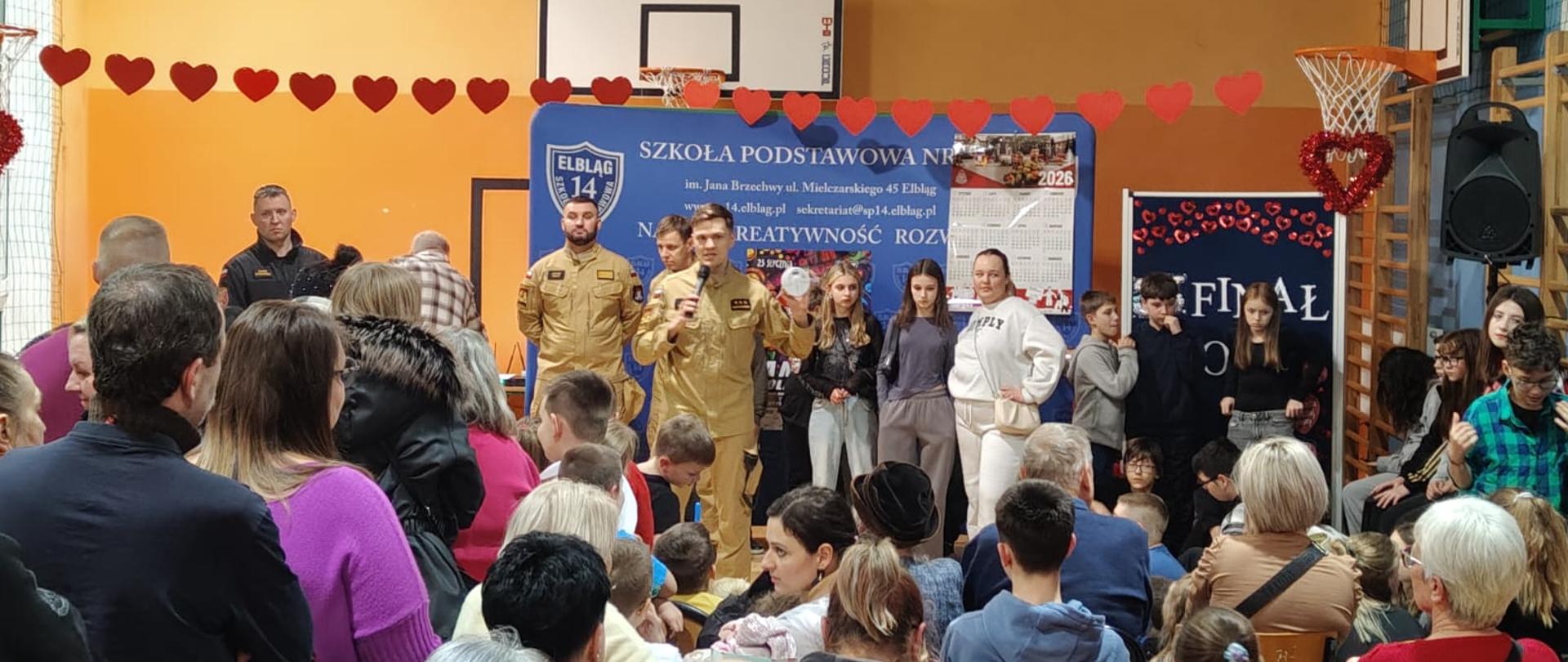 Strażacy w jasnych, piaskowych mundurach stoją pośród młodzieży szkolnej na sali gimnastycznej i opowiadają o zasadach bezpieczeństwa. W tle na ścianie tablica do gry w kosza.