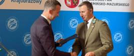 Mężczyzna w garniturze podaje rękę mężczyźnie w mundurze. W tle znajduje się ścianka z napisem „Welski Park Krajobrazowy”.