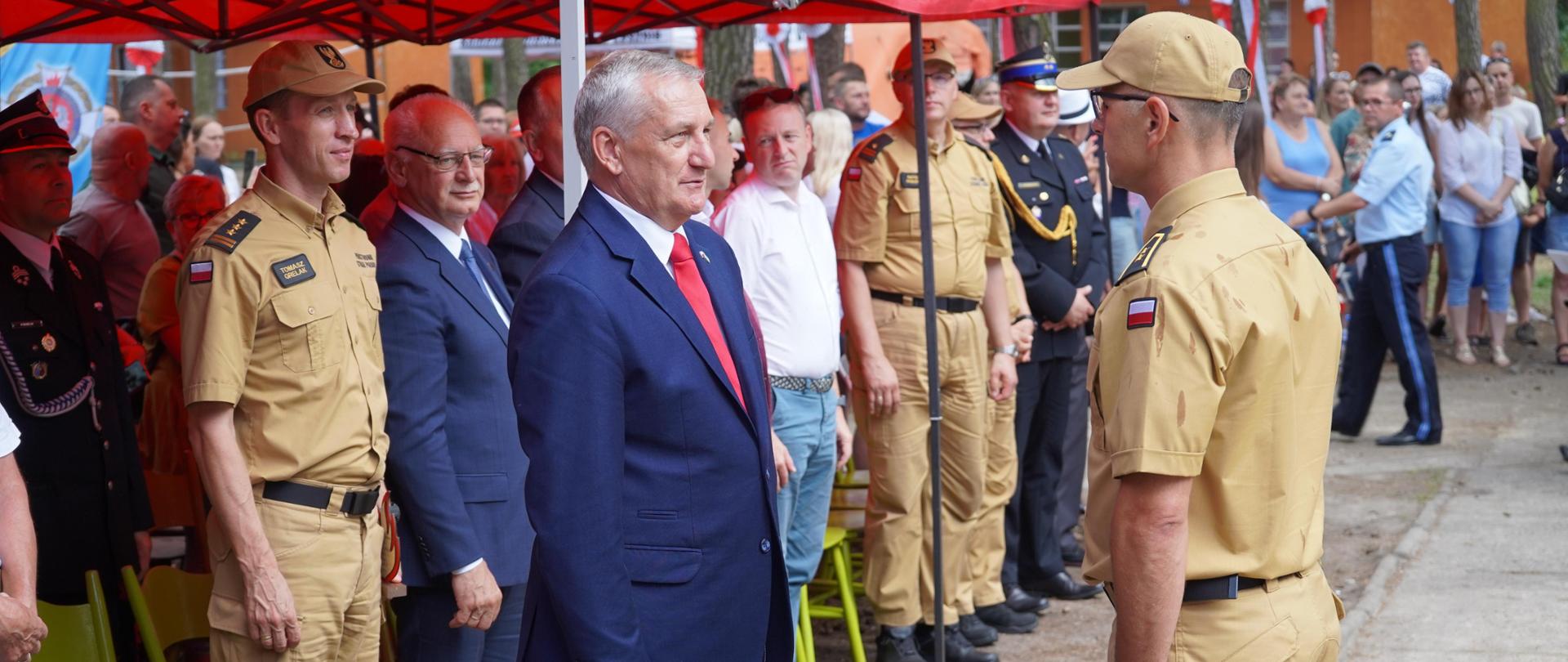 Dowódcę obozu meldującego o rozpoczęciu uroczystości wiceministrowi Spraw Wewnętrznych i Administracji. Za nimi stoją pod namiotami zaproszeni goście.