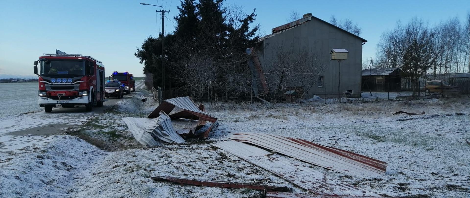 Zdjęcie przedstawia kilka arkuszy zdeformowanej blachy z przykrycia dachowego. Blacha znajduje się na ziemi, na której zalega biała warstwa śniegu. W tle widać zaparkowane na drodze dwa samochody ratowniczo-gaśnicze oraz jeden pojazd operacyjny państwowej straży pożarnej. 