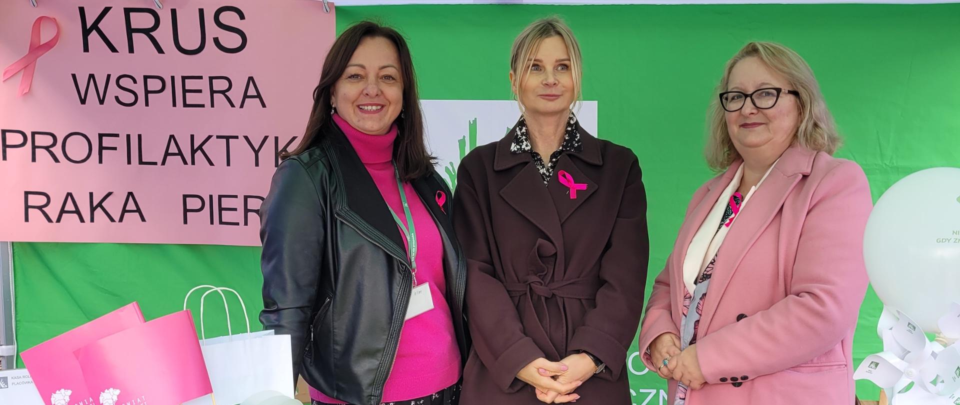 Trzy kobiety stoją przy stoisku informacyjno-prewencyjnym Kasy Rolniczego Ubezpieczenia Społecznego w Myślenicach podczas Marszu Różowej Wstążki. Za nimi widoczny jest baner z napisem „KRUS wspiera profilaktykę raka piersi”. Na stole znajdują się materiały informacyjne, gadżety profilaktyczne i drobne nagrody dla uczestników wydarzenia.