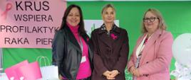 Trzy kobiety stoją przy stoisku informacyjno-prewencyjnym Kasy Rolniczego Ubezpieczenia Społecznego w Myślenicach podczas Marszu Różowej Wstążki. Za nimi widoczny jest baner z napisem „KRUS wspiera profilaktykę raka piersi”. Na stole znajdują się materiały informacyjne, gadżety profilaktyczne i drobne nagrody dla uczestników wydarzenia.