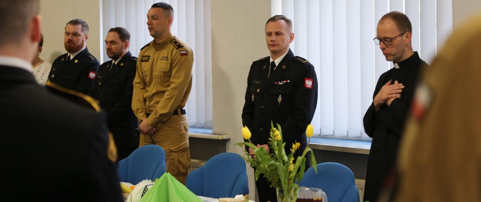 W głębi od lewej stojący przy stole podczas świątecznego spotkania dwaj oficerowie Centralnej Szkoły PSP, aspirant oraz p.o. Komendant Centralnej Szkoły PSP oraz ksiądz