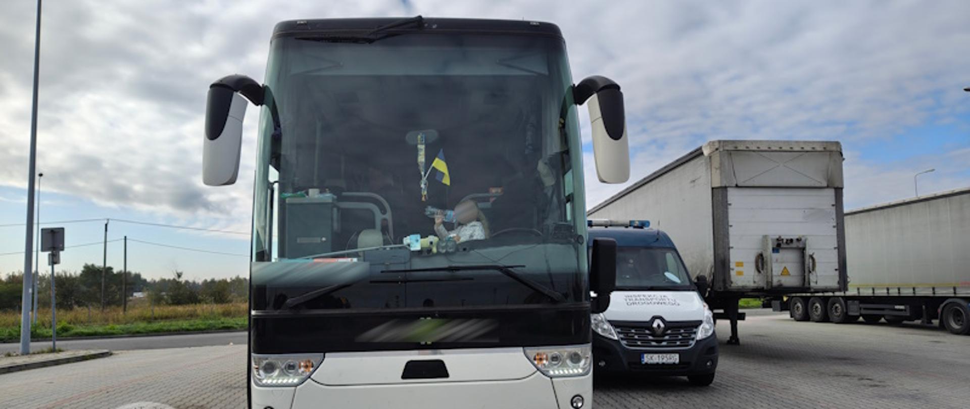 Na zdjęciu widać biały autokar stojący na parkingu drogowym, obok którego zatrzymał się radiowóz inspekcji transportu drogowego. W tle widoczne są inne pojazdy ciężarowe oraz niebo z chmurami. Zdjęcie zostało wykonane podczas kontroli autokaru z Włoch przez polskich inspektorów transportu drogowego.