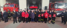 Pomorski komendant wojewódzki Państwowej Straży Pożarnej stoi wspólnie z zaproszonymi gośćmi oraz przedstawicielami Młodzieżowych Drużyn Pożarniczych którzy trzymają czerwone teczki.