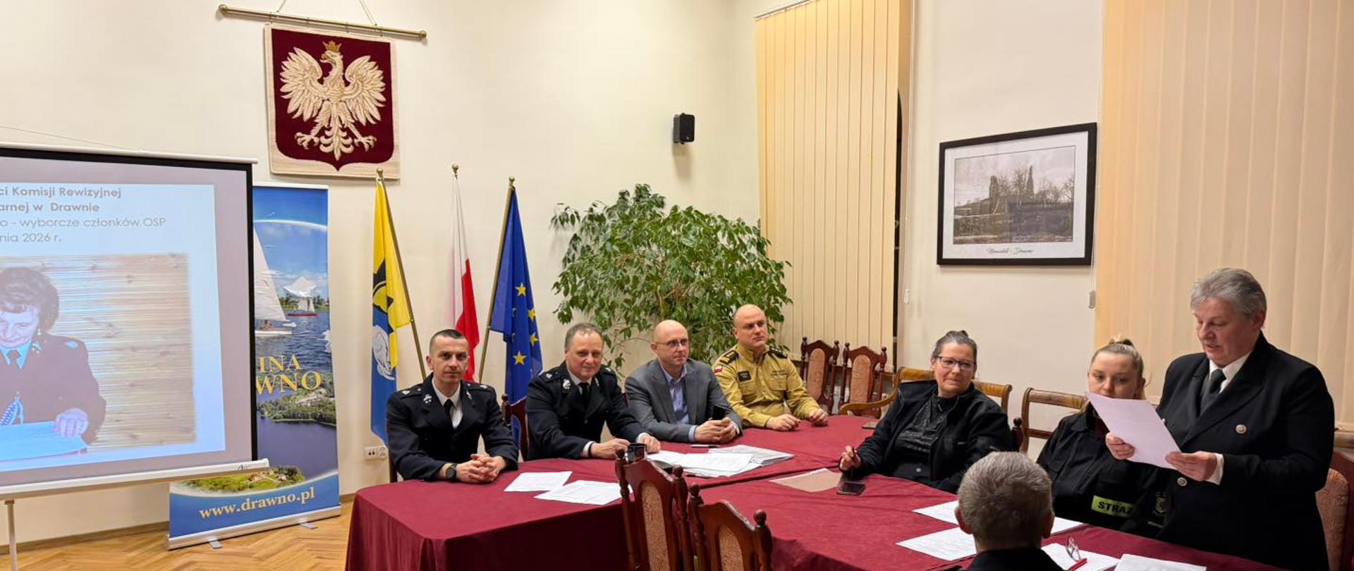 Sala konferencyjna. Przy długich stołach siedzą uczestnicy posiedzenia, część w mundurach strażackich, część w strojach cywilnych. Jedna osoba stoi i czyta dokument. W tle godło Polski, flagi, roleta okienna oraz ekran z prezentacją.