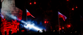 Pomnik podświetlony w barwach flagi Polski pod pomnikiem stoją harcerze oddając honory na masztach wiszą flagi Polski wystrzelono race.