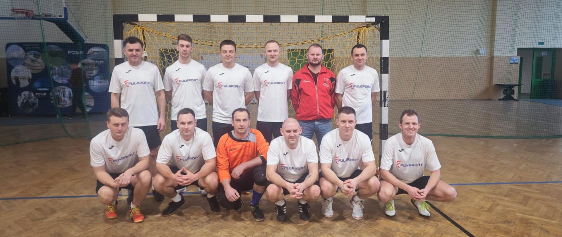 Kolorowa fotografia wykonana w pomieszczeniu. Przedstawia strażaków Komendy Powiatowej PSP w Brzozowie podczas turnieju piłki nożnej. Strażacy w strojach sportowych ustawieni w bramce na sali gimnastycznej.