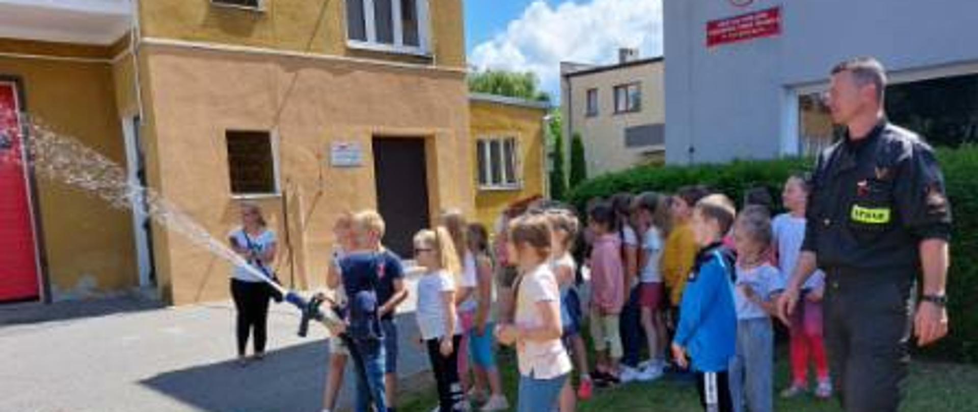 uczniowie pod nadzorem strażaka leją wodę z prądownicy na placu komendy