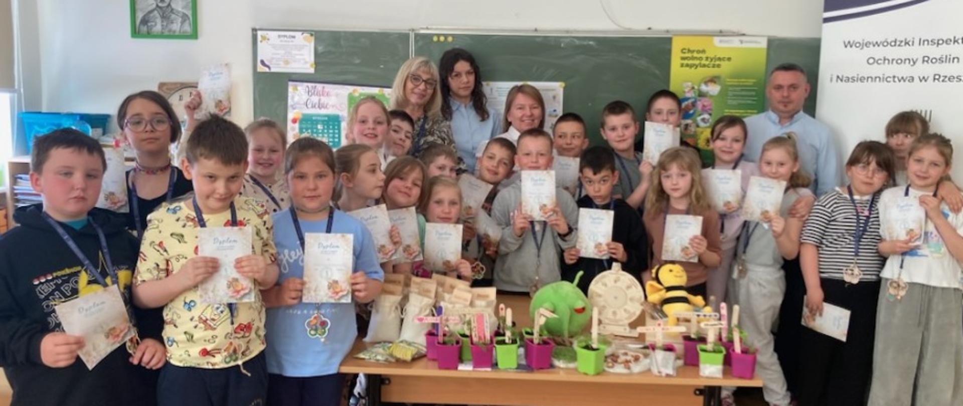 Grupowe zdjęcie w klasie szkolnej przedstawiające kilkanaścioro dzieci wraz z nauczycielami. Dzieci stoją w kilku rzędach, większość trzyma w rękach dyplomy i nosi medale na szyjach. Przed nimi stoi stół z różnymi materiałami edukacyjnymi i małymi doniczkami z roślinami. W tle znajduje się tablica szkolna oraz dekoracje ścienne. Atmosfera jest uroczysta i radosna, jak po zakończeniu projektu lub konkursu.