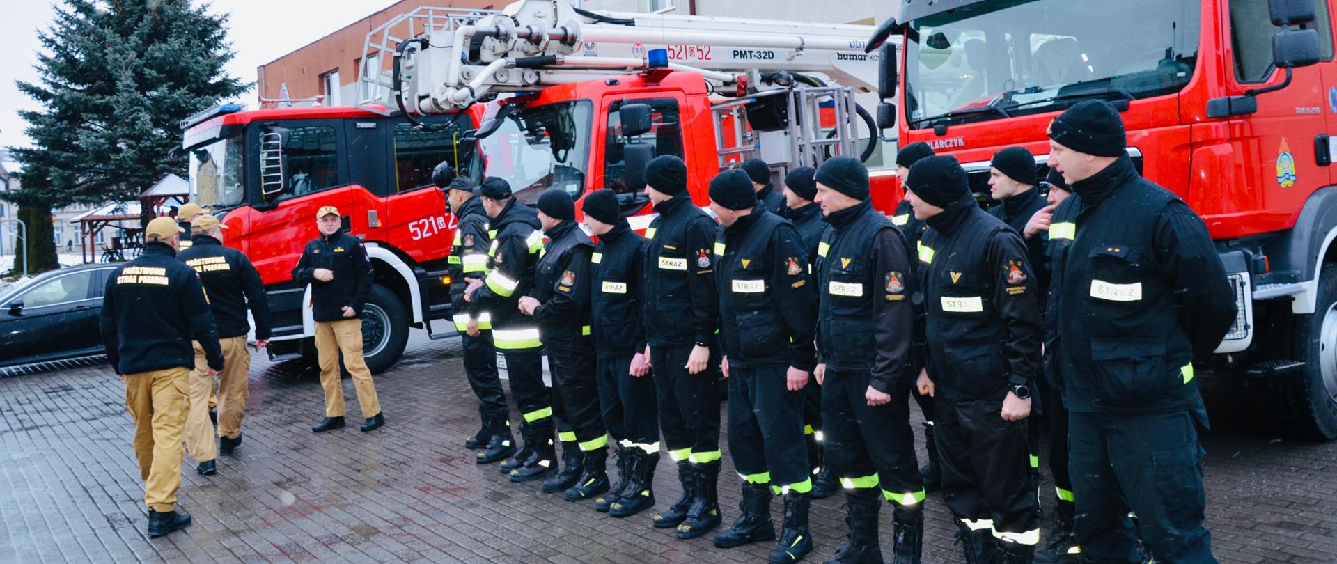 Zdjęcie przedstawia strażaków ustawionych w szeregu przed samochodami jednostki. Kadra kierownicza KW wizytuje jednostkę, w tle budynki garaży JRG.