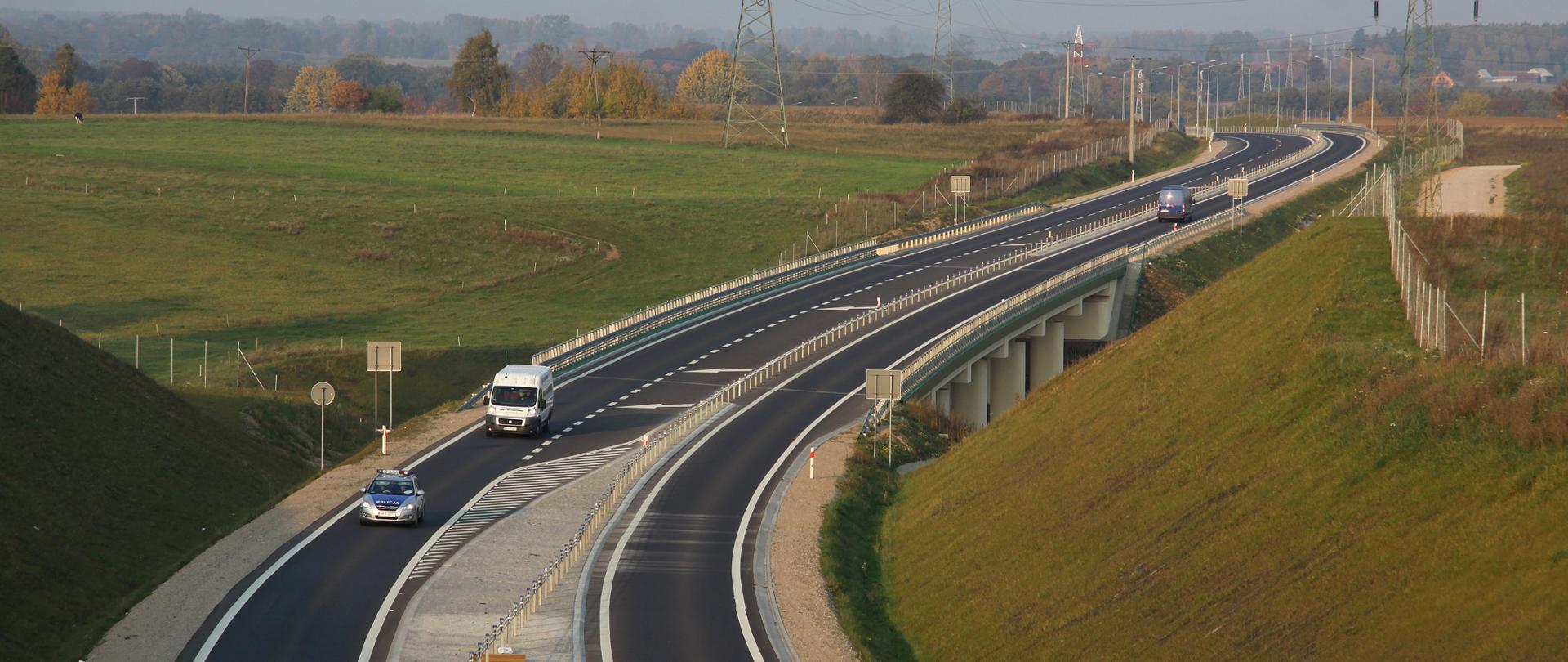 Droga przebiegająca przez tereny zielone, łąki. Na drodze widoczne są trzy samochody, w oddali znajdują się maszty linii energetycznej. Na horyzoncie znajdują się liczne drzewa.