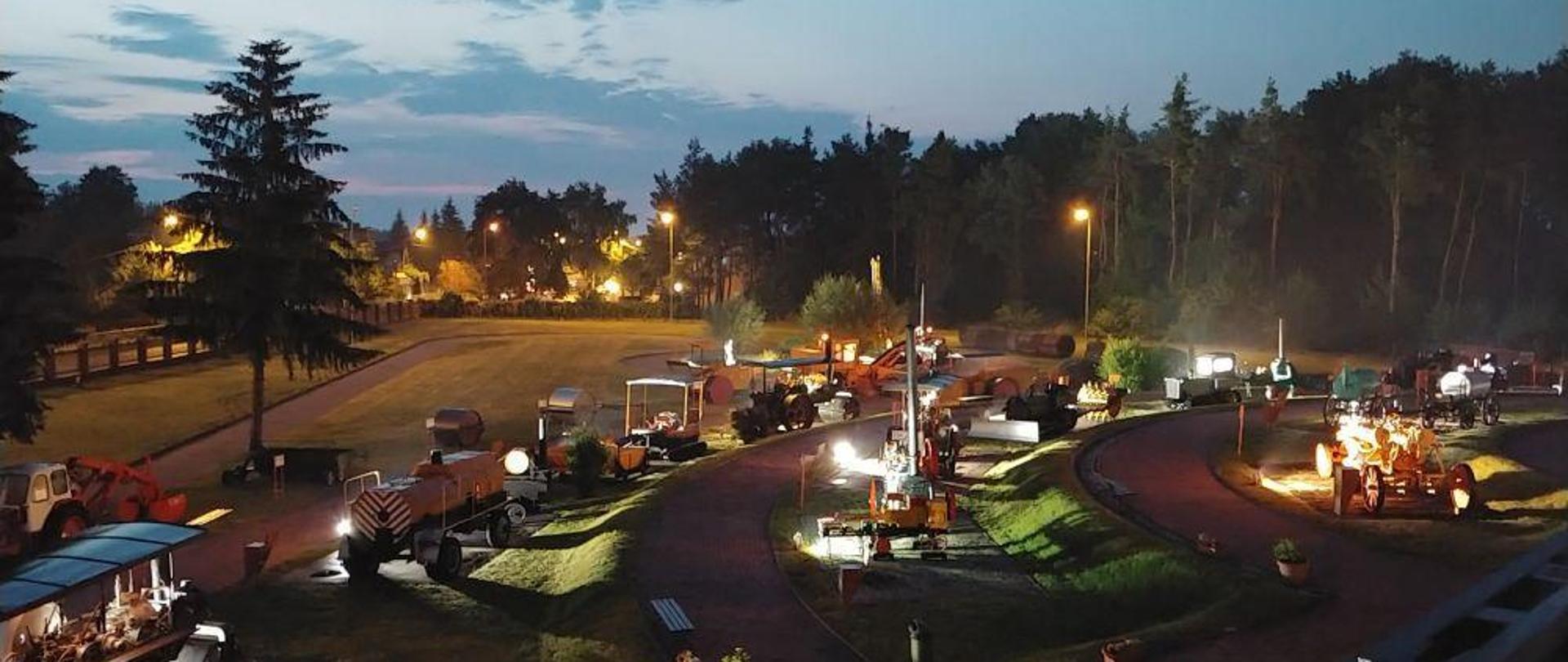 Nocne zdjęcie terenu wokół budynku muzeum. Widoczne eksponaty muzealne, drzewa i oświetlenie z lamp
