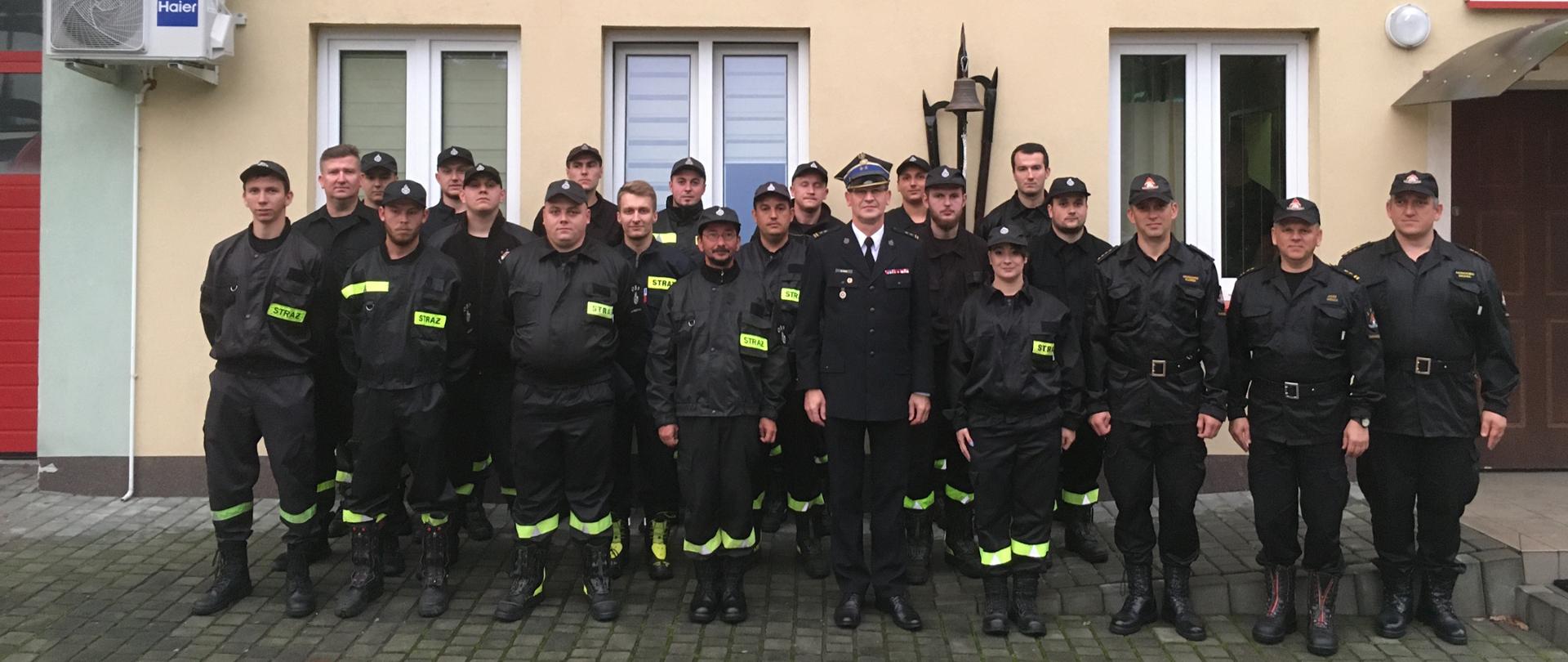 Przed budynkiem ustawiona grupa strażaków w umundurowaniu