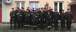 Przed budynkiem ustawiona grupa strażaków w umundurowaniu.