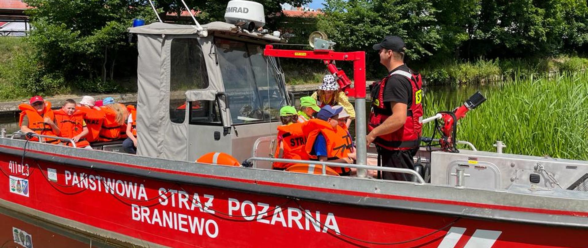 Grupa dzieci w pomarańczowych kamizelkach na czerwonej łodzi strażackiej. W tle zielone drzewa.