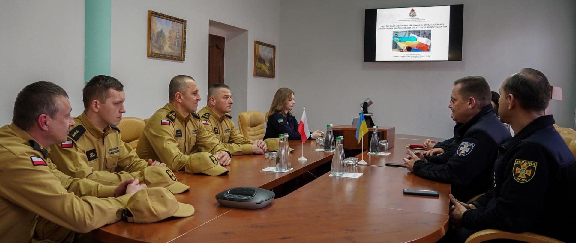 Zdjęcie zrobione wewnątrz pomieszczenia podczas spotkania polskich ukraińskich strażaków. Przy stole konferencyjnym siedzą polscy i ukraińscy strażacy uczestniczący w spotkaniu.