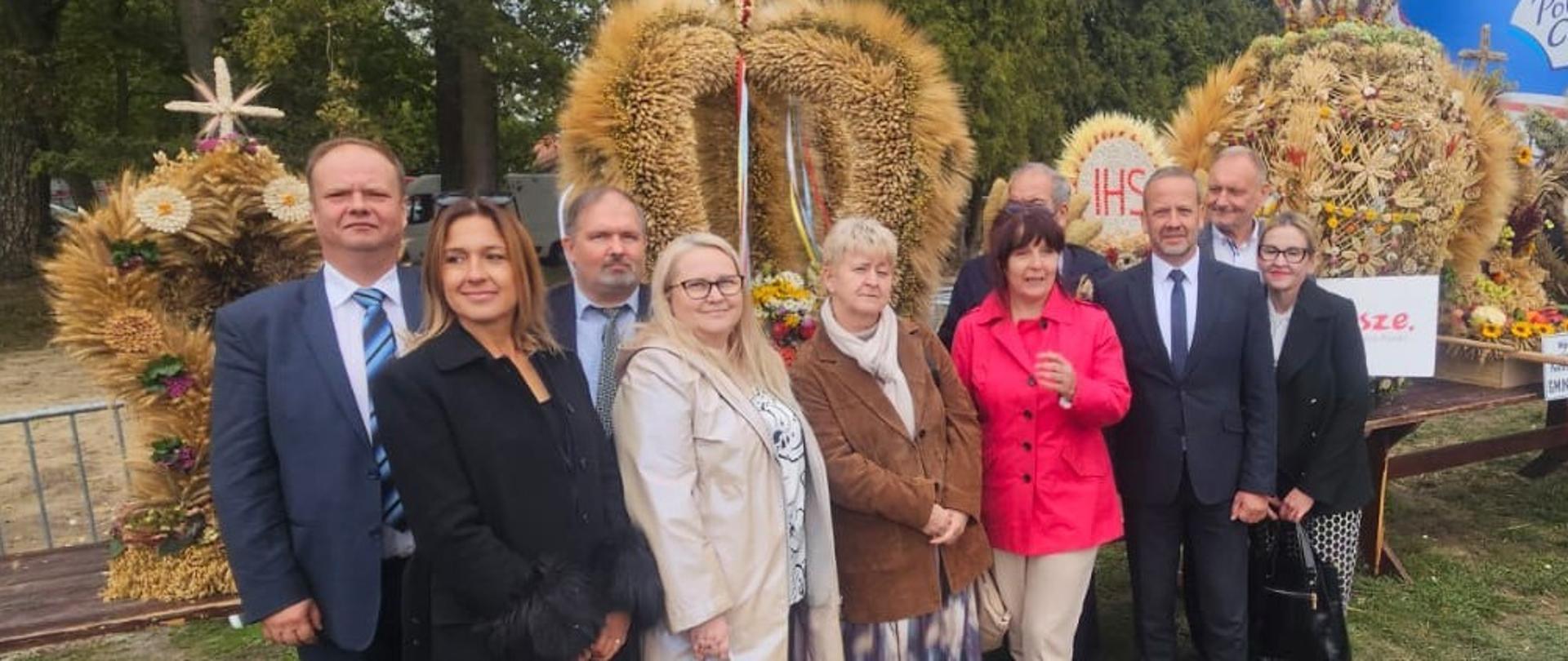 Grupa osób, kobiet i mężczyzn pozująca na tle wieńców dożynkowych