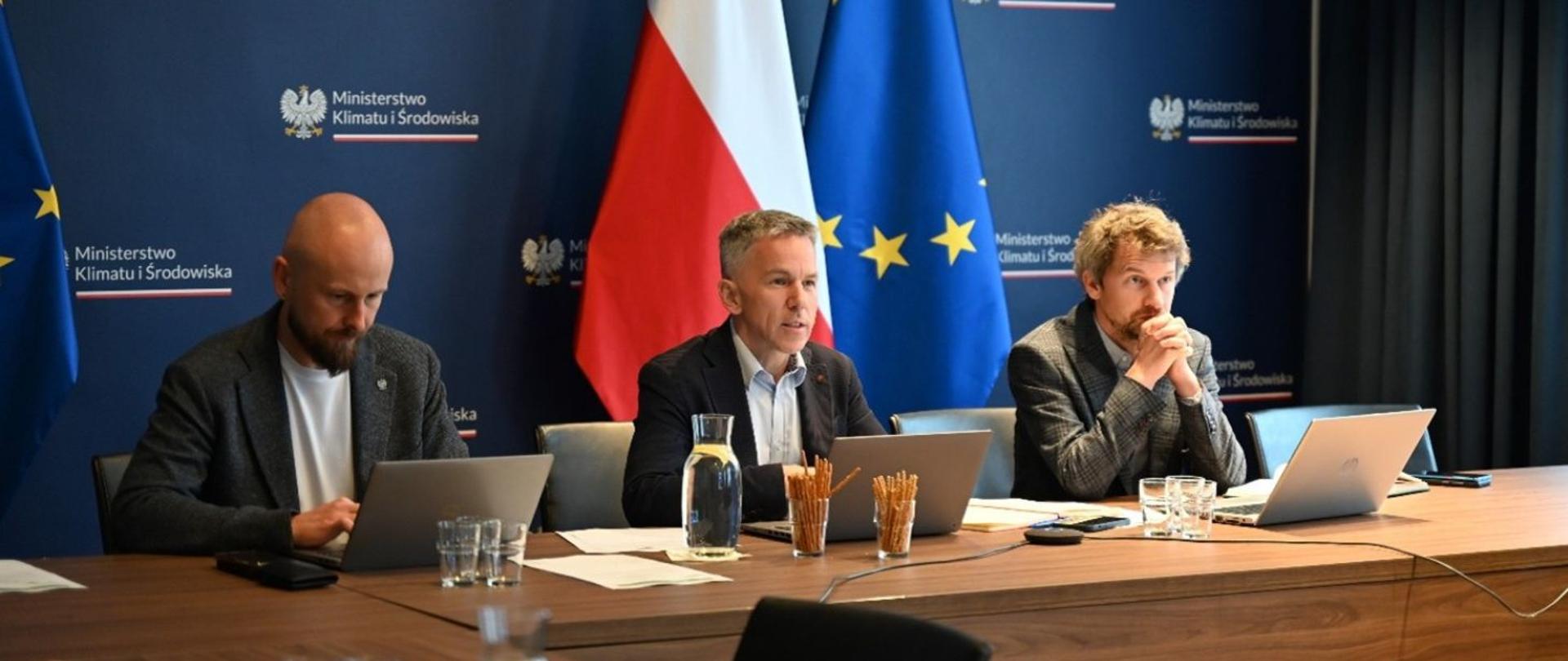 Na zdjęciu widać 3 mężczyzn siedzących za biurkiem przed komputerami. Na biurku stoi również karafka z wodą, szklanki oraz przekąski w formie paluszków. Leżą również dokumenty. W tle widać ściankę z logotypem Ministerstwa Klimatu i Środowiska oraz flagi Polski oraz Unii Europejskiej.