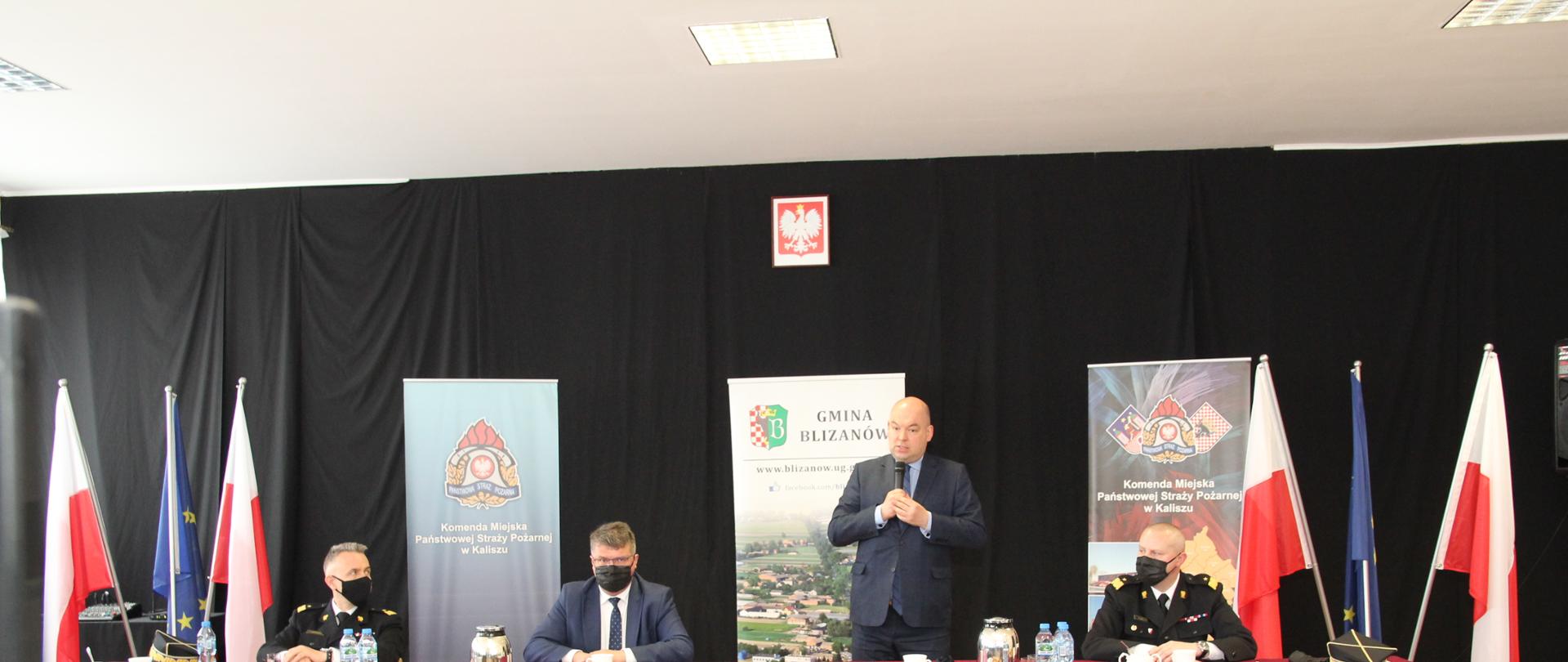 przy dużym stole siedzi trzech mężczyzn, w środku jeden stoi trzymając mikrofon, w tle banery , flagi państwowe oraz czarna ściana