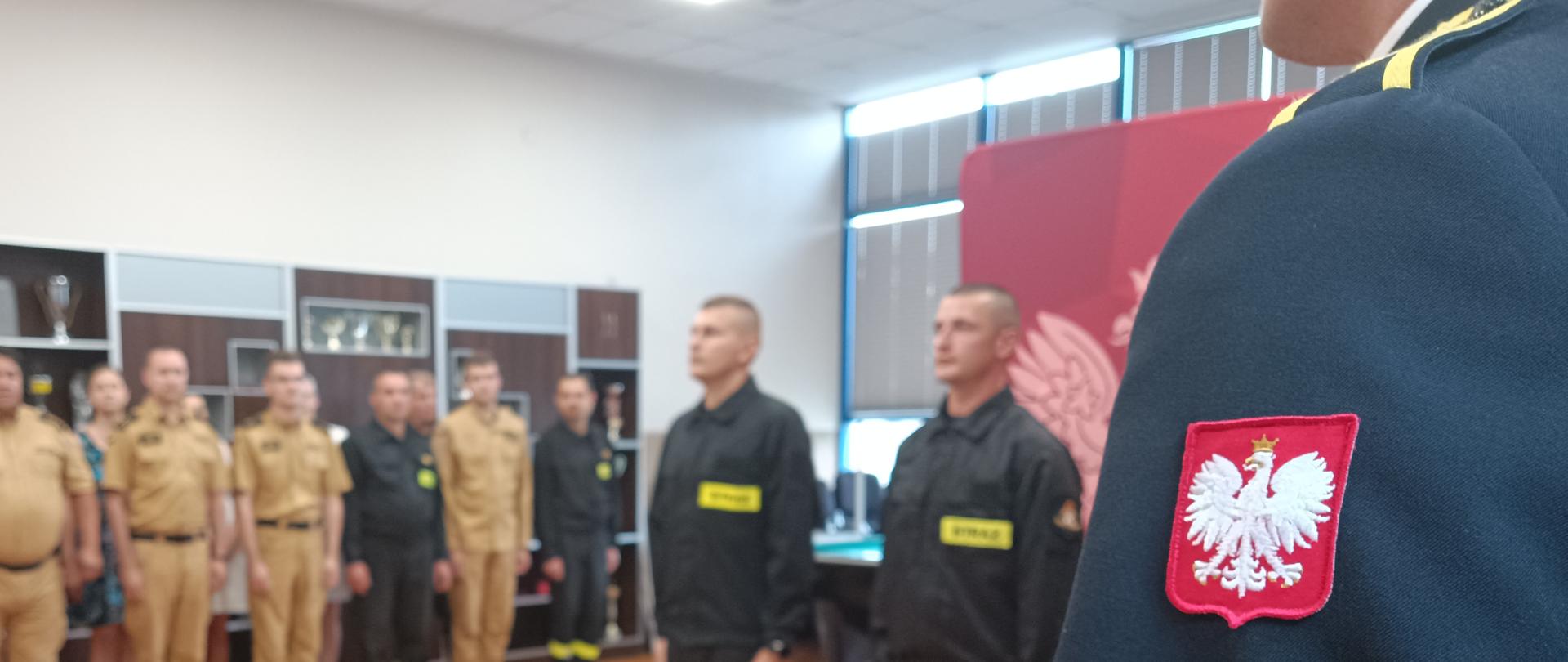 Na zdjęciu w tle pracownicy KP PSP w Pabianicach, po prawej stronie nowo przyjęci do służby przygotowawczej strażacy. Na pierwszym planie funkcjonariusz wchodzący w skład pocztu sztandarowego.