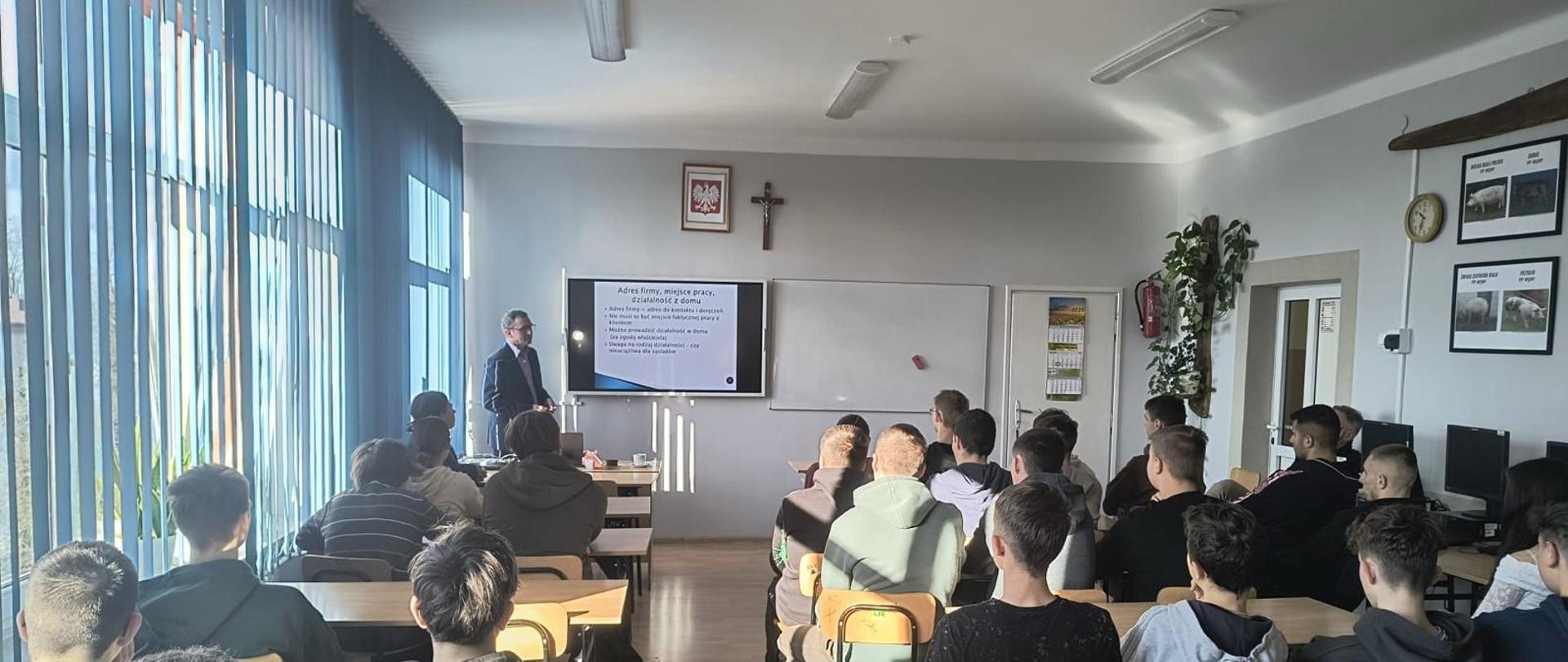 W jasnej sali lekcyjnej siedzi grupa uczniów zwrócona w stronę osoby stojącej przy ekranie, na którym wyświetlana jest prezentacja. Do pomieszczenia wpada światło dzienne przez rząd niebieskich żaluzji, a na ścianach widoczne są dekoracje i tablica.
