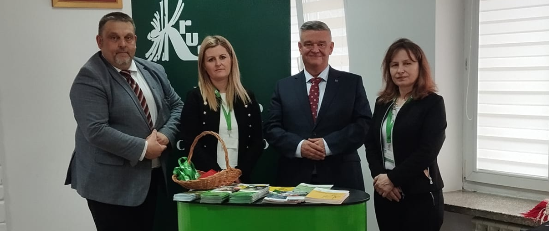 Stoisko informacyjne KRUS w holu Komendy Państwowej Straży Pożarnej w Proszowicach. Na pierwszym planie trybunka reklamowa z logo KRUS z materiałami prewencyjnymi. Przy stoisku dwie kobiety i dwóch mężczyzn pozują do zdjęcia. Za nimi stoi rollup z logo KRUS.