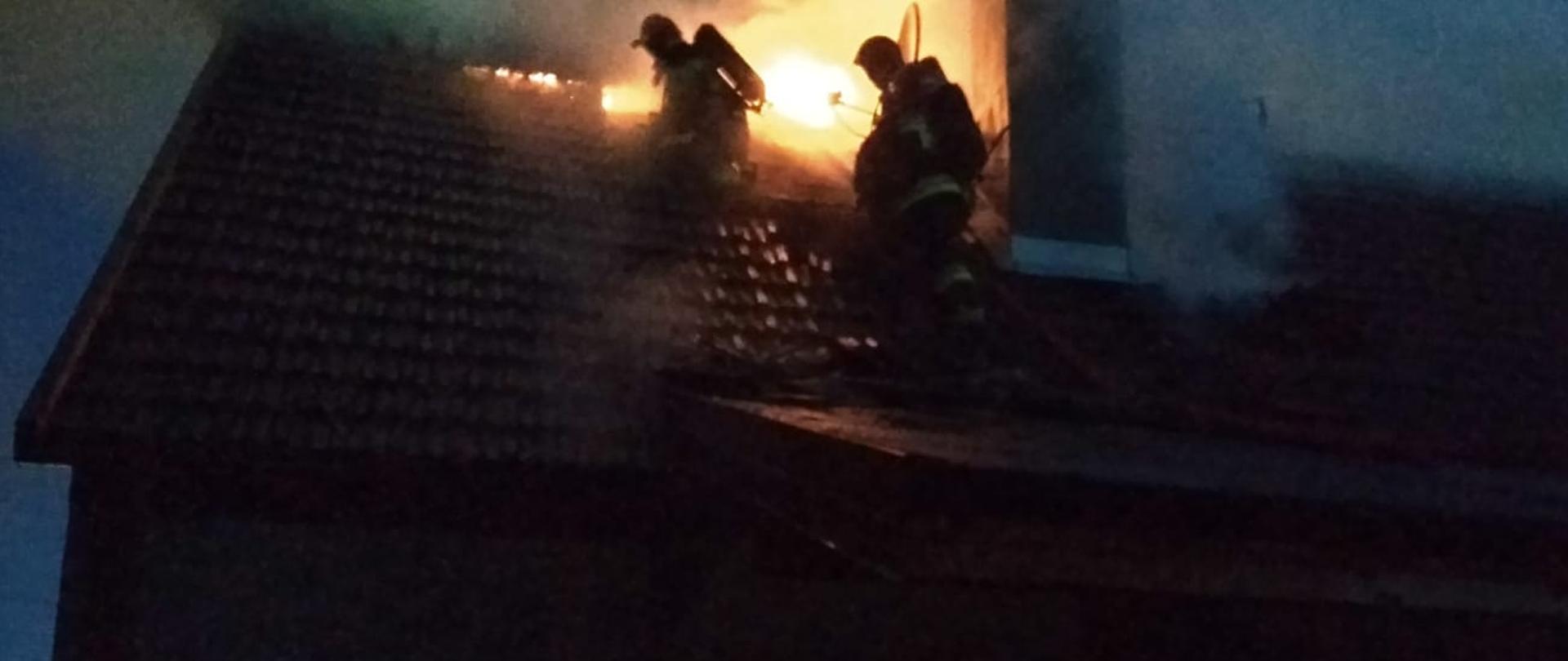 Zdjęcie przedstawia wydobywające się płomienie z dachu budynku mieszkalnego jednorodzinnego oraz dwóch strażaków na dachu domu podejmujących działania gaśnicze w celu ugaszenia pożaru. Z prawej strony zdjęcia widać również trzeciego strażaka