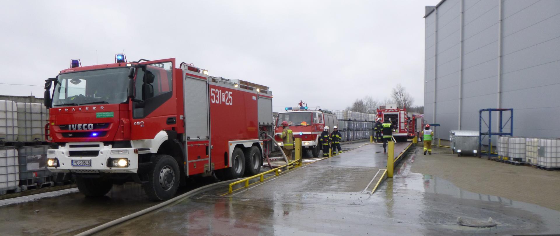 Wozy strażackie obok hali i chodzący strażacy.