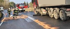 Strażacy OSP zbierają z jezdni wyciekłe płyny eksploatacyjne z pojazdu ciężarowego. W tle widać samochód pożarniczy i samochód ciężarowy stojący na poboczu jezdni.