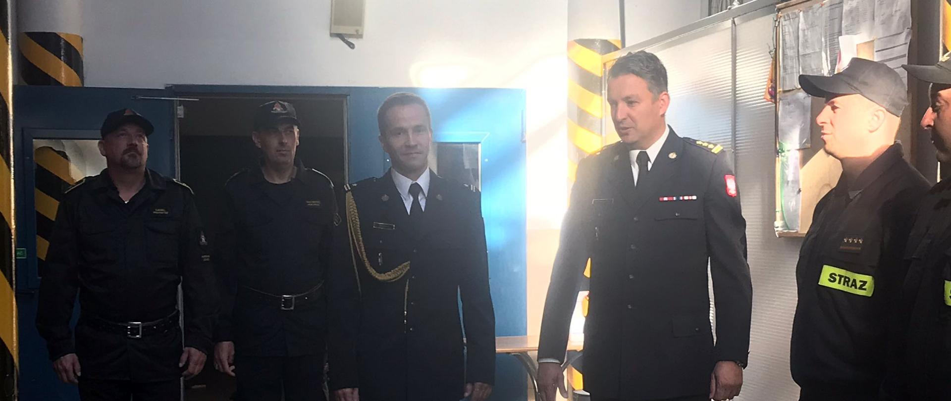 Strażacy i Komendanci stoją w garażu JRG podczas powitania mł. bryg. Dariusza Kosiora