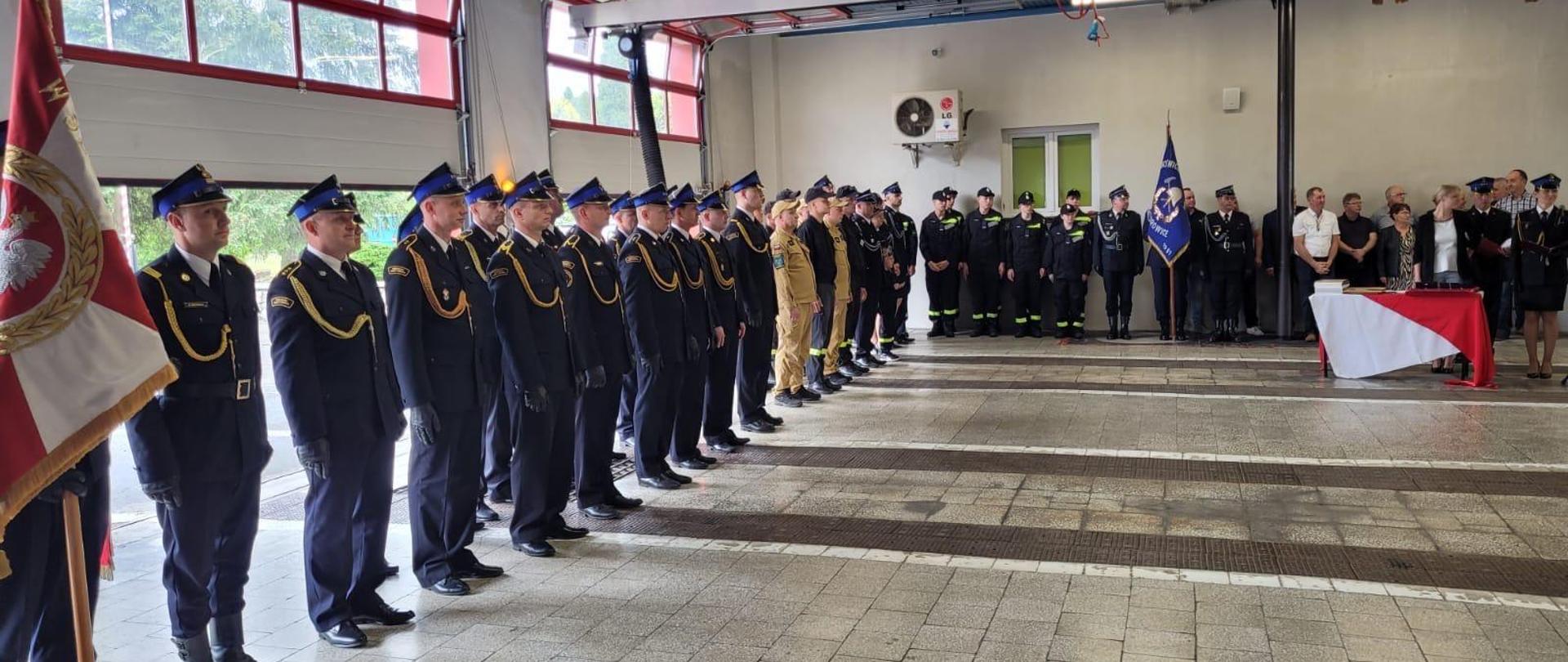 Na zdjęciu znajdują się strażacy w mundurach wyjściowych ustawieni w szyku. W tle widać pomieszczenia garażowe