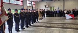 Na zdjęciu znajdują się strażacy w mundurach wyjściowych ustawieni w szyku. W tle widać pomieszczenia garażowe