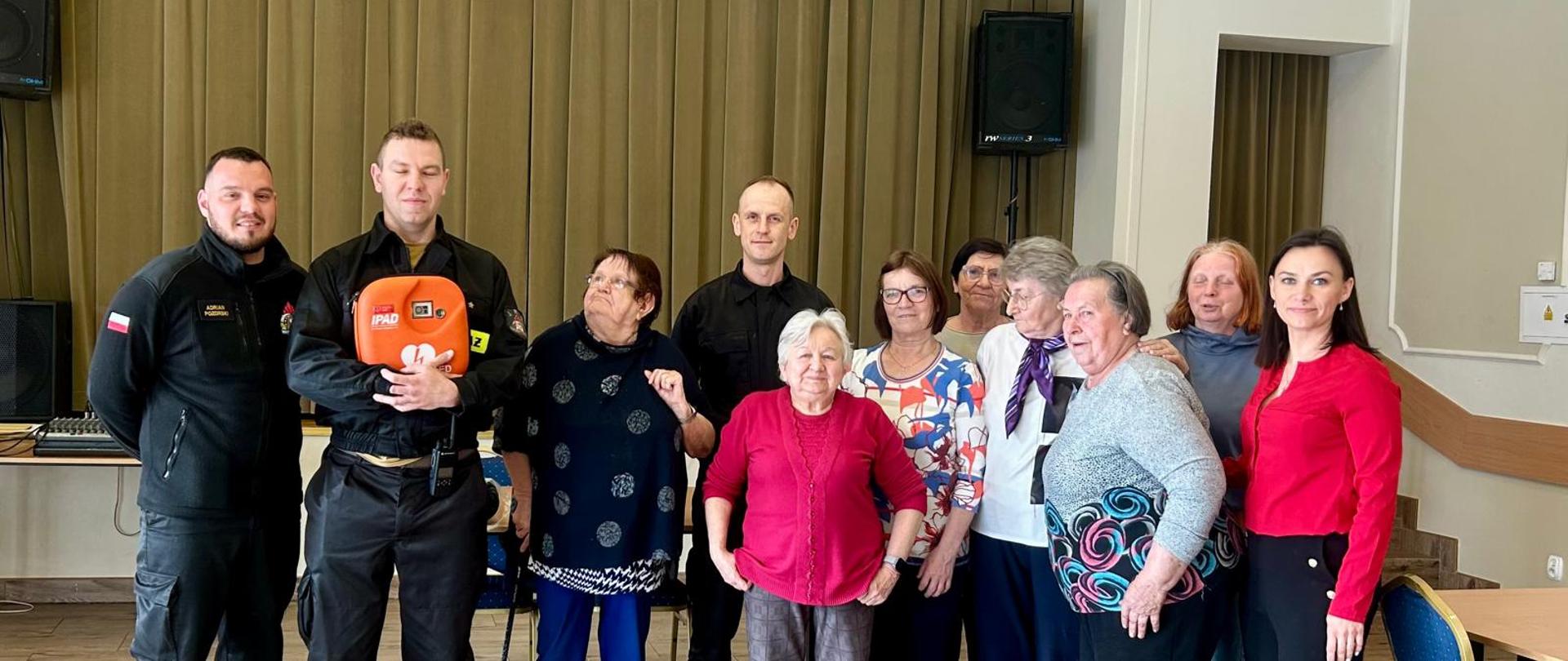 trzech strażaków w ubraniach koszarowych oraz zgromadzeni goście pozują do zdjęcia na auli klubu seniora.