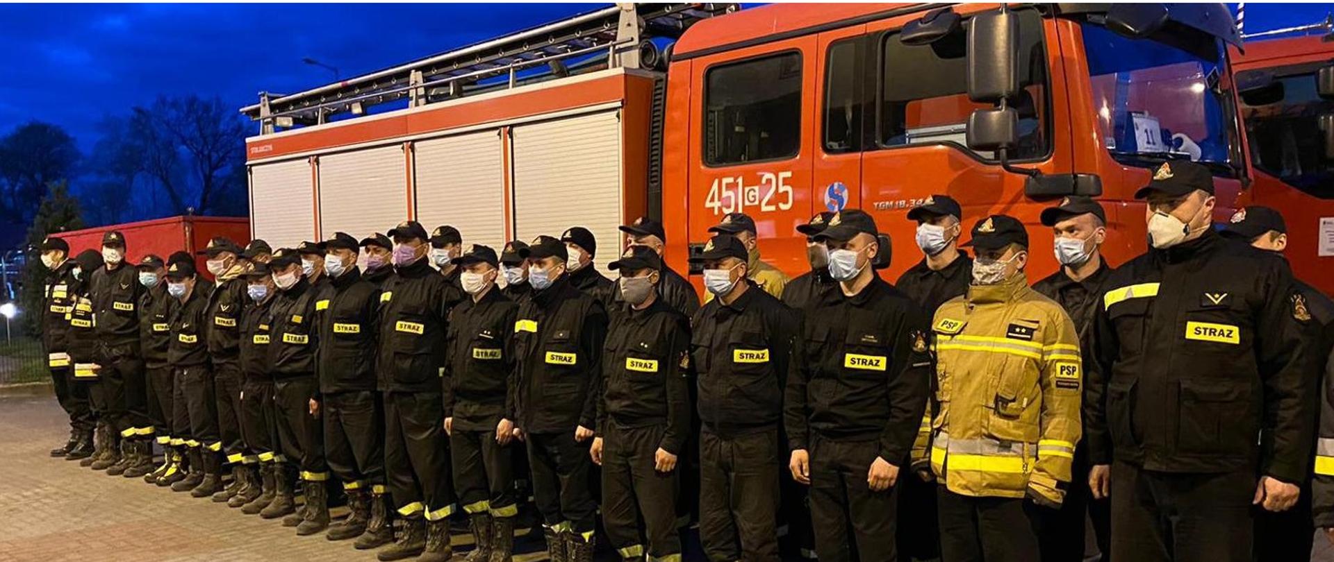 Strażacy z maseczkami zakrywającymi nos i twarz, stojący w dwuszeregu, na tle ciężkiego samochodu gaśniczego