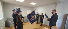 Zbiórka strażaków w jasnym pomieszczeniu. Po lewej stronie stoi trzech strażaków w mundurach wyjściowych. Środkowy trzyma granatowy sztandar komendy. W środku dwoje funkcjonariuszy (kobieta i mężczyzna) ubranych w mundury koszarowe składa ślubowanie. Po prawej komendant w mundurze wyjściowym odczytuje rotę ślubowania z granatowej teczki. Wszyscy zgromadzeni mają twarze zasłonięte maseczkami. W tle wyposażenie gabinetu: stół, fotele.