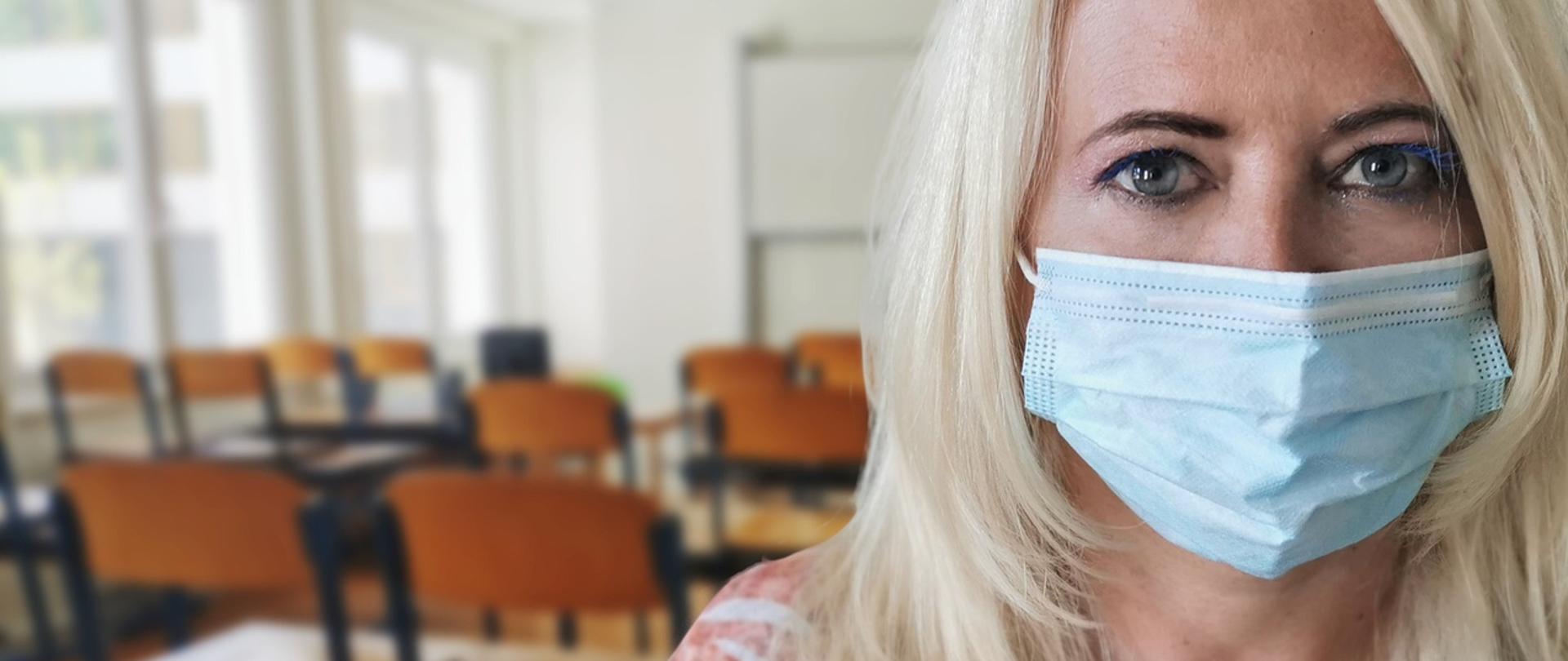 nauczycielka w maseczce jednorazowej na twarzy, a za nią puste krzesła przy ławkach w szkolnej klasie.