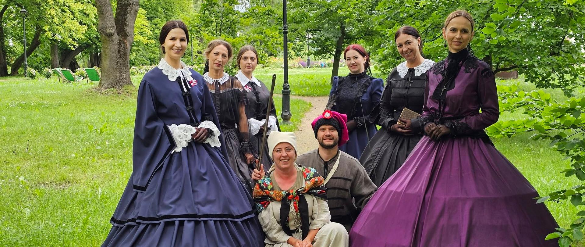 Grupa osób w historycznych kostiumach pozuje do zdjęcia na parkowej alejce. Uczestniczki stoją w długich sukniach stylizowanych na dawne stroje, a dwie osoby siedzą z przodu w ludowych i historyzujących przebraniach. W tle widoczna jest zieleń parku, drzewa i alejka spacerowa.