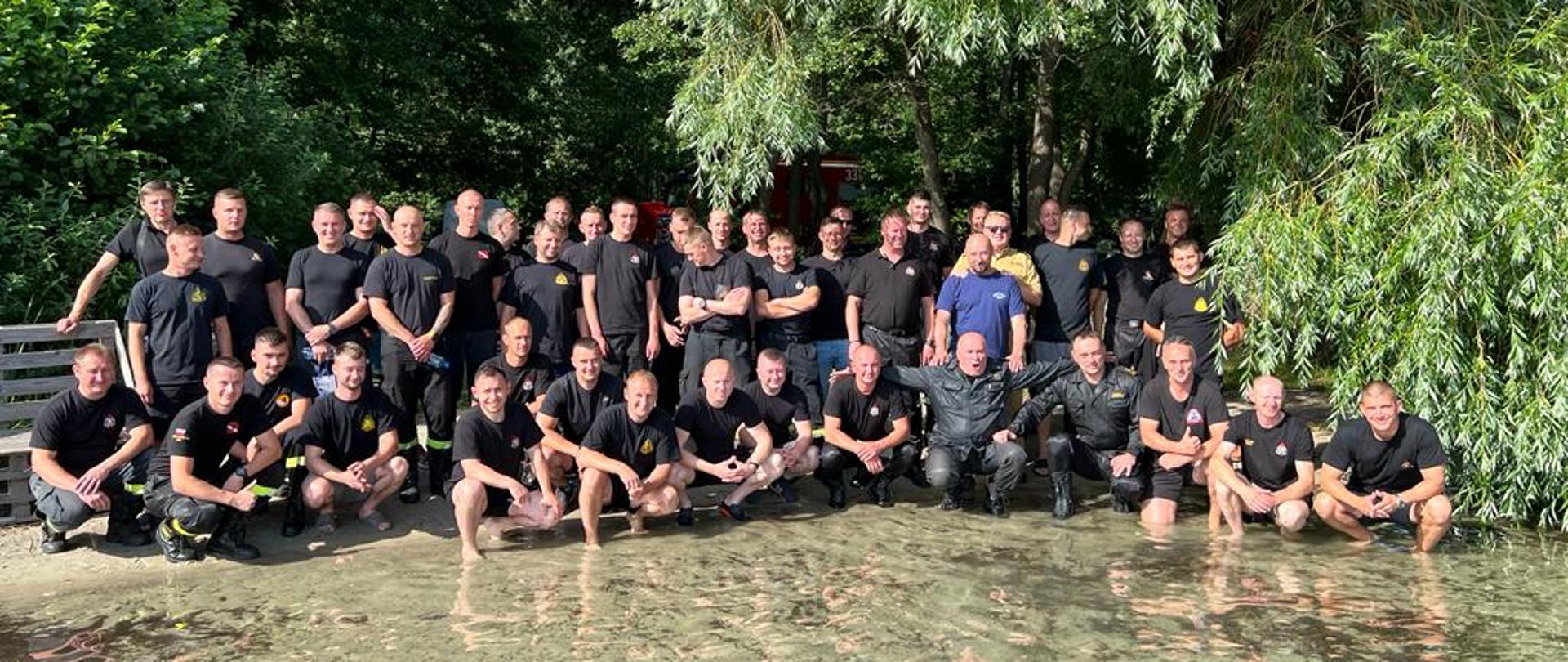 grupa strażaków stojąca na brzegu jeziora pozują do zdjęcia w tle drzewa
