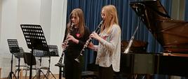 Zdjęcie przedstawia dwie klarnecistki grające na swych instrumentach z nut stojących przed nimi na pulpicie. Z tyłu po prawej stronie otwarty czarny fortepian. Z lewej strony pulpity i krzesła tworzące nieład. Uczennice ubrane są w czarne spodnie, jedna w białą druga w czarną koszulę. Włosy maja długie opadające na ramiona, na piersiach przyklejona naklejka z serduszkiem czerwonym WOŚP-u.