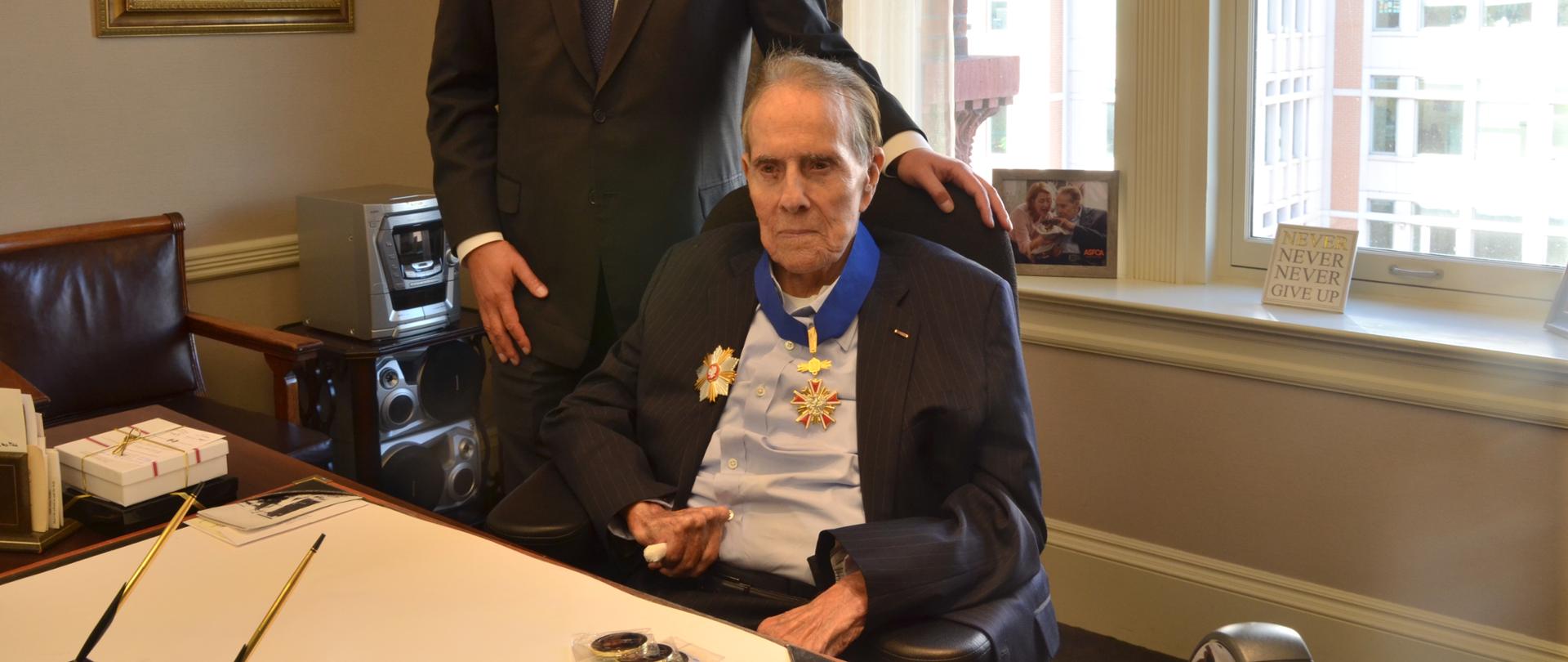 Udekorowany odznaczeniem senator Robert Dole siedzi na krześle. Obok stoi Ambasador Piotr Wilczek