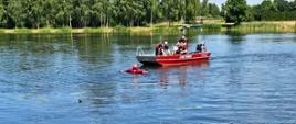 Strażacy na łodzi ratunkowej pomagają osobie w wodzie trzymającej koło ratunkowe na tle zieleni i spokojnej tafli jeziora.