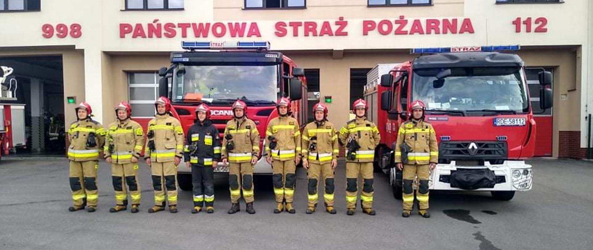 Strażacy w mundurach używanych podczas działań ratowniczo-gaśniczych oddają hołd Powstańcom Warszawskim. Stoją na placu wewnętrznym na tle samochodów pożarniczych, które mają włączone sygnały świetlne