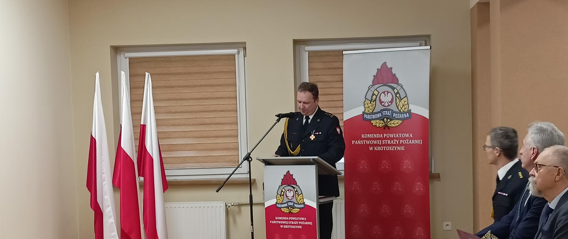 Zdjęcie przedstawia komendanta powiatowego PSP podczas omawiania materiału analitycznego. Funkcjonariusz stoi przy mównicy, w tle stojak z flagami Polski i baner Komendy Powiatowej PSP.