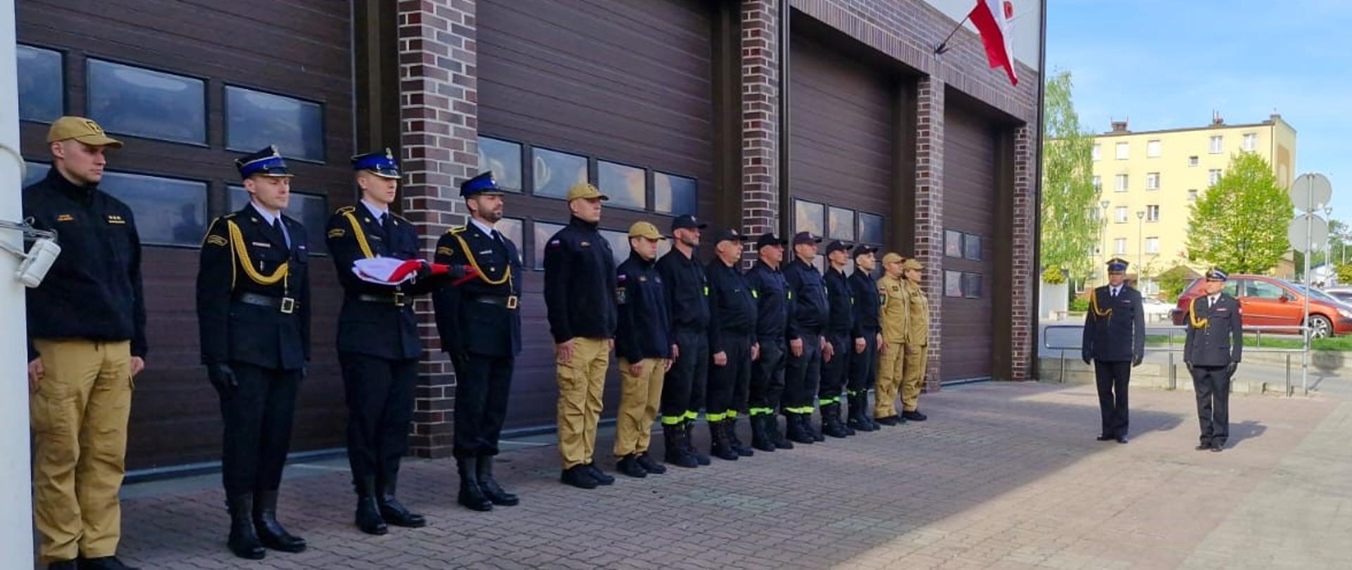 
Zdjęcie przedstawia uroczystość przed budynkiem Komendy Powiatowej Państwowej Straży Pożarnej w Ostrowi Mazowieckiej, odbywającą się w słoneczny dzień. Na pierwszym planie, na brukowanym placu, widoczny jest rząd około piętnastu strażaków stojących na baczność. Trzech z nich, znajdujących się na lewym krańcu rzędu, jest w galowych mundurach, a środkowy z nich trzyma złożoną polską flagę. Pozostali strażacy są ubrani w ciemne, bojowe mundury z żółtymi elementami lub w piaskowe mundury polowe.
Po prawej stronie zdjęcia, nieco z boku, stoi dwóch strażaków w galowych mundurach. W tle znajduje się budynek straży pożarnej z dużymi, brązowymi bramami garażowymi. Nad bramami widnieje czerwony napis "STRAŻ POŻARNA", a nad jedną z nich wisi mała polska flaga. W tle widoczne są również drzewa i inne budynki oraz częściowo błękitne niebo.