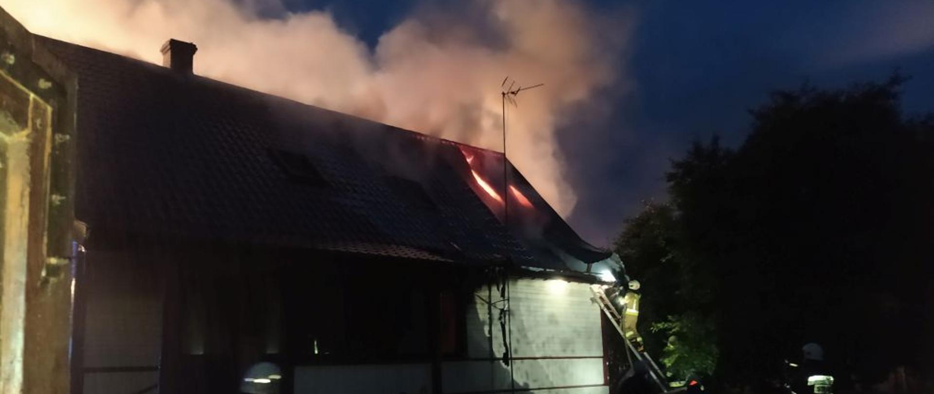 W nocy. Budynek jednorodzinny biały. Przez blaszane pokrycie wydobywają się płomienie. Jeden ze strażaków stoi na drabinie opartej o dach budynku. Trzech strażaków w ciemnych mundurach bojowych prowadzi akcję gaśniczą.