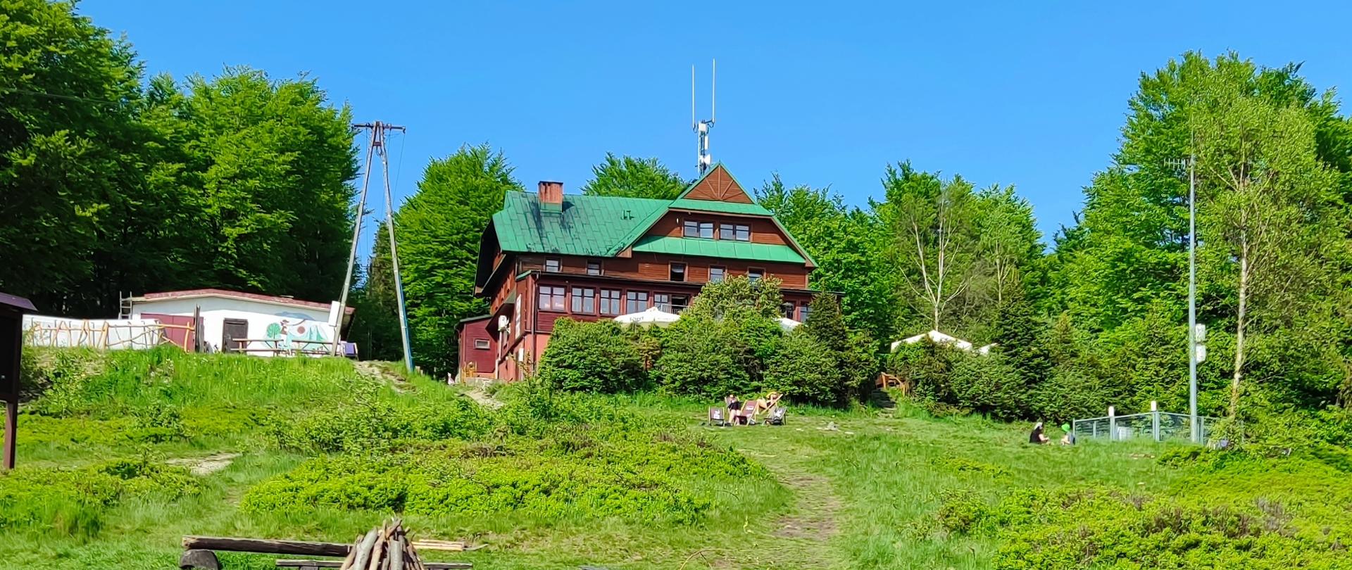 Młode osoby wypoczywające na trawie, w słońcu, w tle budynek schroniska