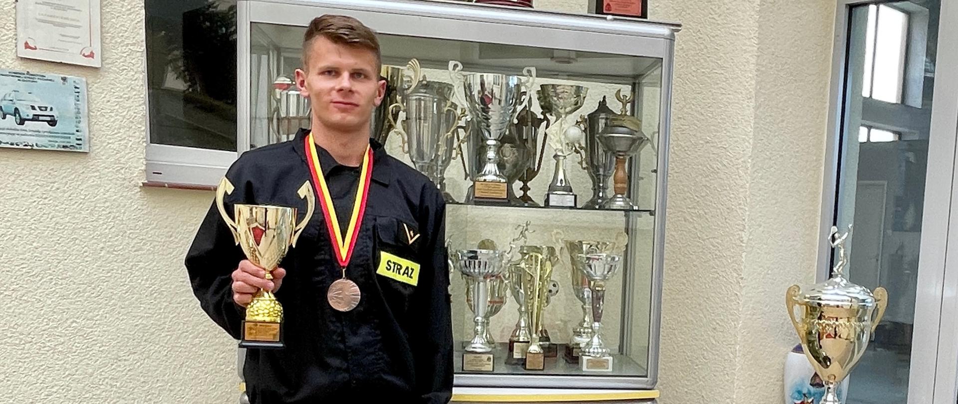 strażak w mundurze z wiszącym na szyi medalem trzymający puchar 