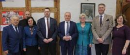 Zdjęcie zbiorowe, minister stoi w otoczeniu kilku elegancko ubranych osób na tle białej ściany ozdobionej kilkoma zbitymi na krzyż deskami.