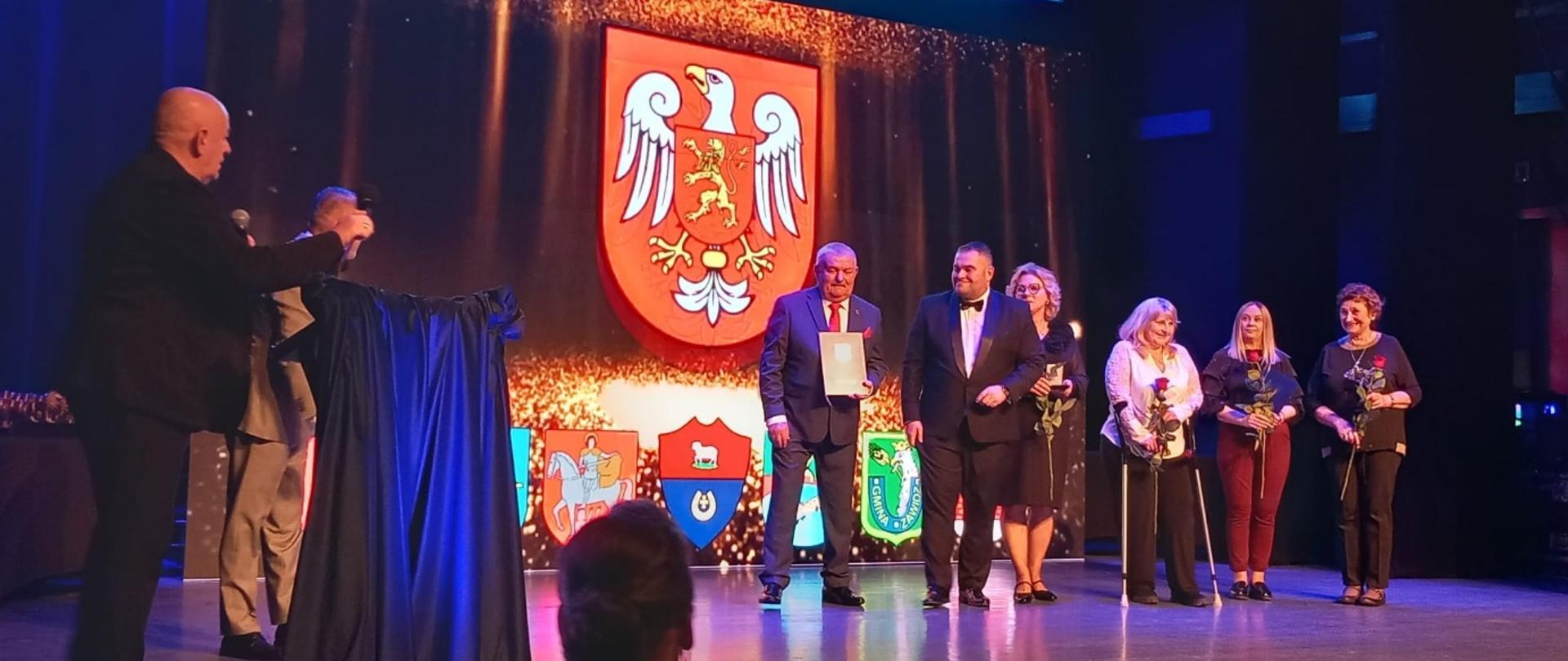 Na scenie podczas oficjalnej uroczystości kilka osób stoi w rzędzie, a jeden z mężczyzn trzyma dyplom lub wyróżnienie. W tle widoczny jest duży herb oraz grafika z napisem „2026”, a pod nim znajdują się herby gmin. Część uczestników trzyma kwiaty, a wydarzenie odbywa się w oświetlonej sali widowiskowej.